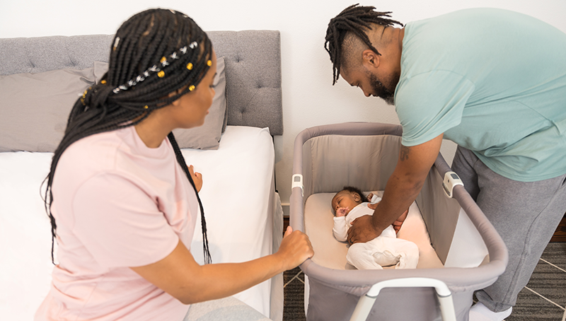 A Father and Mother puting their baby on their back to sleep in a crib. 