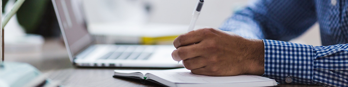 A man takes notes next to an open laptop.